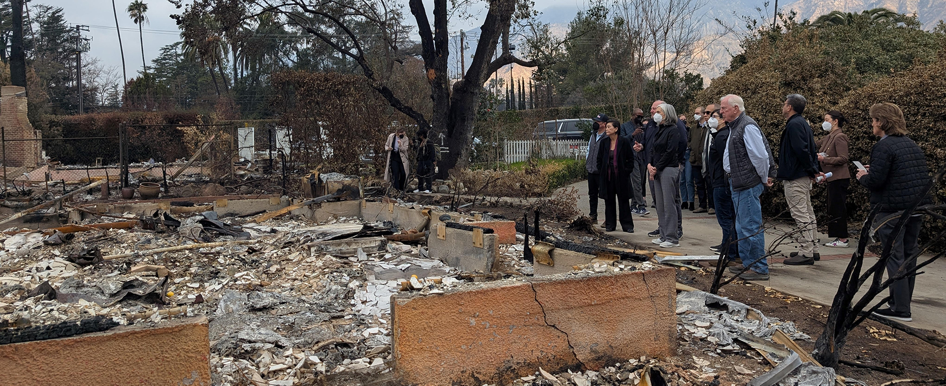 A group of people, including President Joe Biden, touring the aftermath of a wildfire with destroyed structures and debris in the foreground and mountains partially obscured by haze in the background.